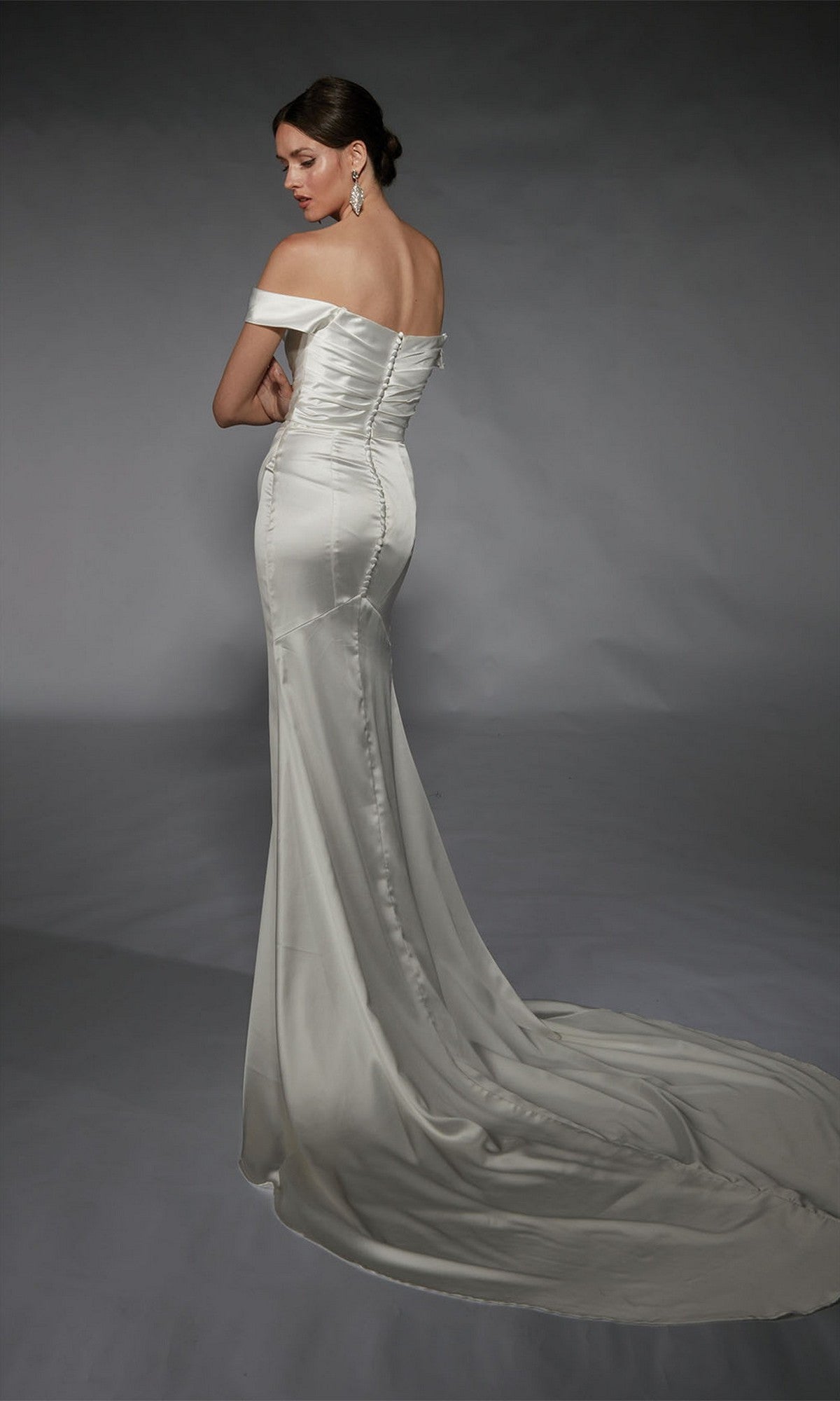 A woman faces away against a plain backdrop, wearing the Alyce 7106 long white dress—an off-the-shoulder satin gown with a fitted silhouette, long train, and detachable overskirt for extra elegance.